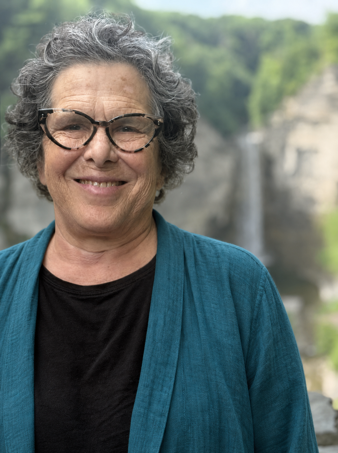 Amy Glin smiling outdoors in a teal jacket with a waterfall in the distance